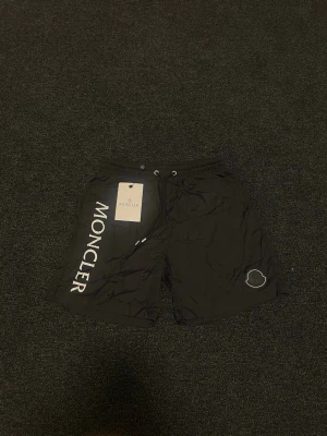 Svarta badbyxor från Moncler - Snygga svarta badbyxor från Moncler med vit logga längs ena benet och en liten Moncler-symbol på andra benet. Klassisk passform med dragsko i midjan och snabbtorkande material. Perfekta för stranden eller poolen.