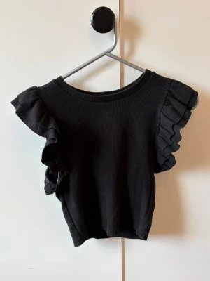 Svart topp med volangärmar - Snygg svart ribbad topp med volangdetaljer på ärmarna. Toppen har rund halsringning och är tillverkad i ett mjukt, stretchigt material som sitter bekvämt. Perfekt för dig som vill ha en enkel men ändå trendig look.