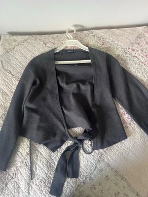Mörkgrå omlottkofta med knytband - Snygg mörkgrå omlottkofta med långa ärmar och breda knytband i midjan. Koftan har en öppen front utan knappar och är tillverkad i ett mjukt, tunt material som känns skönt mot huden. Perfekt att slänga över en topp för en avslappnad look. Är i mycket bra skick har använts två gånger! Men inget täcken på användning 
