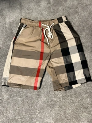 Burbery shorts - Snygga shorts med klassiskt rutigt mönster i beige, svart, vitt och rött. Shortsen har elastisk midja med vit snörning och sidofickor. Perfekta för sommaren och ger en trendig look. Passar S/m