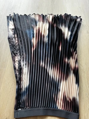 Plisserad kjol med batikmönster H&M - Säljer en snygg plisserad kjol från H&M i storlek S. Kjolen har ett coolt batikmönster i svart, beige och blå toner. Tillverkad i ett glansigt syntetmaterial som ger ett lyxigt intryck. Perfekt för dig som vill sticka ut lite extra!