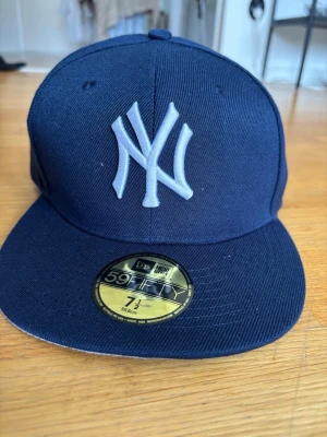 Mörkblå New Era Yankees keps - Säljer en mörkblå New Era 59FIFTY keps med New York Yankees-logga broderad i vitt framtill. Klassisk flat brim och strukturerad passform. Kepsen är i modellen fitted och har en guldfärgad etikett på skärmen. Perfekt för dig som gillar streetwear och sportig stil.