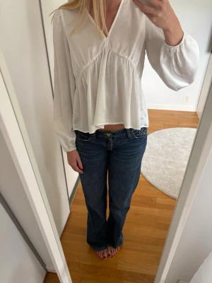 Vit v-ringad blus med volang - Snygg vit blus från AWARE med v-ringning och volangdetaljer under bysten. Blusen har långa ärmar med lätt puff och är tillverkad i ett tunt, flowy material som ger en luftig känsla. Den ser lite genomskinlig ut på galgen men inte i verkligheten. 