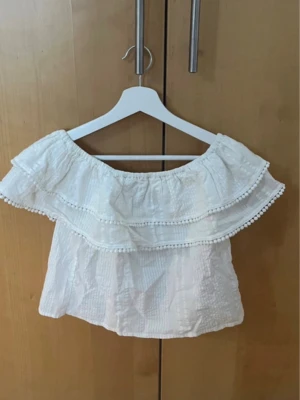 Vit offshoulder blus med volang - Superfin vit blus med offshoulder-modell och dubbla volanger upptill. Blusen har små bollar längs volangkanten och är tillverkad i ett lätt, krinklat tyg. Perfekt för en somrig och avslappnad look.