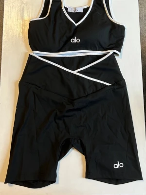 Svart set från Alo med vita detaljer - Snyggt svart tvådelat set från Alo med sport-bh och cykelbyxor. Setet har vita kontrastkanter och logga framtill på både överdel och underdel. Materialet är stretchigt och följsamt, perfekt för träning eller chill. Överdelen har v-ringning och byxorna har hög midja med omlott-detalj.