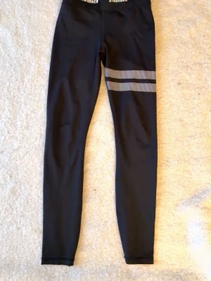 Svarta leggings från Stronger - Snygga svarta leggings från Stronger med två vita randiga detaljer på ena benet. Tillverkade i stretchigt material med 75% återvunnet tyg. Perfekta för träning eller chill. Hög midja och smal passform.