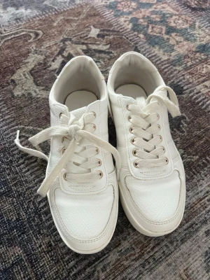 Vita klassiska sneakers i skinn - Snygga vita sneakers med lågt skaft och klassisk design. Skorna har vita snören, guldiga öljetter och är tillverkade i ett slätt och lätt texturerat skinnmaterial. Perfekta för en clean och stilren look.