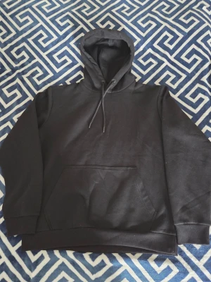 Svart hoodie med huva och magficka - En klassisk svart hoodie med stor huva och dragsnören. Hoodien har en rymlig magficka framtill och ribbade muddar vid ärmslut och nederkant. Tillverkad i mjukt material, perfekt för en avslappnad stil.