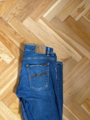 Blå jeans från Nudie - Ett par klassiska blå jeans från Nudie med smal passform och fem fickor. Jeansen har en snygg tvätt och den ikoniska Nudie-sömmen på bakfickan. Perfekta för en avslappnad och trendig look.