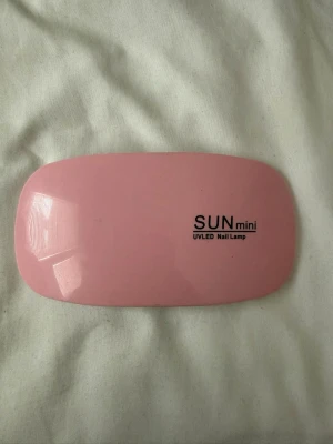 Rosa UV LED nagellampa SUN mini - Säljer en kompakt och smidig UV LED nagellampa i modellen SUN mini. Lampan är rosa på ovansidan och vit undertill, med sex LED-lampor och utfällbara ben. Perfekt för att härda gelélack hemma. Tillverkad i plast.