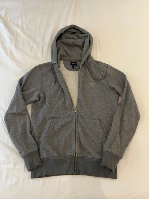 Grå zip hoodie från GANT - Klassisk grå hoodie från GANT med dragkedja, huva med dragsko och två fickor framtill. Liten GANT-logga broderad på bröstet. Tillverkad i mjukt bomullsmaterial som är perfekt för en avslappnad stil.