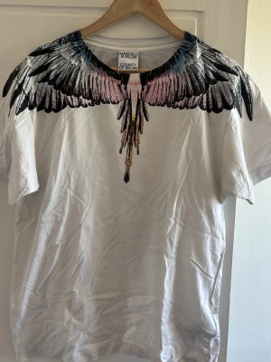 Vit t-shirt med färgglatt fjädertryck - Säljer en vit t-shirt från Marcelo Burlon med ett ikoniskt fjädermönster i svart, rosa, blått och gult runt halsen. T-shirten har rund hals och korta ärmar. Perfekt för dig som vill sticka ut med en unik och trendig design. Har släppt färg från den 