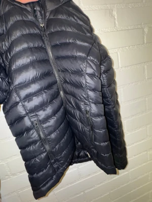 Svart dunjacka  - Snygg svart dunjacka från Everest med quiltad design och två praktiska fickor med dragkedja framtill. En liten reva på armen men man ser den inte när den väl används. Perfekt för hösten eller vintern. Storlek 146-152 så skulle säga xxs eller xs
