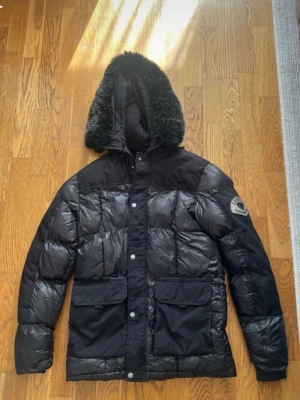 Svart dunjacka med huva - Snygg svart dunjacka med stor huva och fuskpälsdetalj. Jackan har två stora fickor framtill, quiltad design och tryckknappar. Perfekt för kalla vinterdagar och har en patch på ärmen för extra stil.