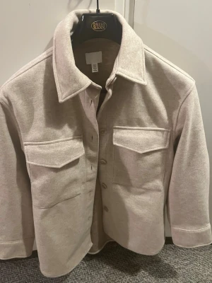 Beige overshirt med stora fickor - Stilren beige overshirt med klassisk krage och stora bröstfickor med lock. Skjortan har knäppning framtill och är gjord i ett mjukt, lite tjockare tyg som ger en avslappnad look. Perfekt att slänga över en t-shirt för en trendig lager-på-lager-stil.