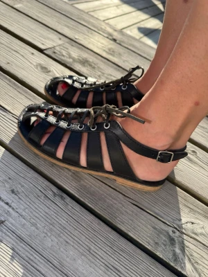 Svarta gladiatorsandaler i läder Swedish hasbeens - Säljer ett par svarta gladiatorsandaler från Swedish hasbeens med snörning framtill och justerbart spänne vid ankeln. Sandalerna har öppen tå och sula i naturfärgat material. Perfekta för sommaren och ger en cool vibe till din outfit. Använda ett fåtal gånger. 