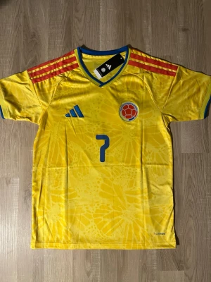 Colombia landslagströja Luis Díaz #7 - Gul Colombia fotbollströja med blå detaljer, röd axelrand och nummer 7. På bröstet finns landslagets emblem och på ryggen står det 'Luis Díaz' samt stort nummer 7. Tillverkad i lätt och ventilerande material. HELT NY!