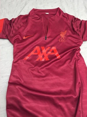 Liverpool tränings-tracksuit röd Nike - Går att köpa enskilt Snygg röd tränings-tshirt från Liverpool FC med orangea detaljer och logga på bröstet. Kort dragkedja framtill, korta ärmar och tryckt AXA-logo på magen. Tillverkad i ett lätt och snabbtorkande funktionsmaterial, perfekt för träning 