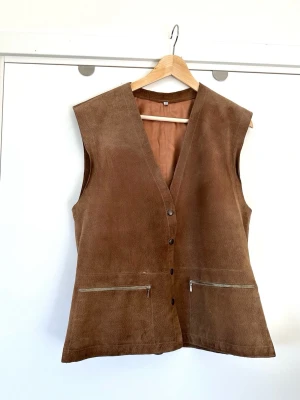 Brun mockaväst med dragkedjefickor - Stilren brun väst i 100% äkta mjuk mocka / skinn / läder i storlek 40. Tidlös modell med knappar framtill, två fickor med dragkedja fram och justerbara spännen för bästa passform i ryggen. Insidan är fodrad i ett matchande brunt tyg. Lika snygg att bära som den är eller stylad i lager på lager över skjorta, blus, klänning eller annat. Mycket fint skick!