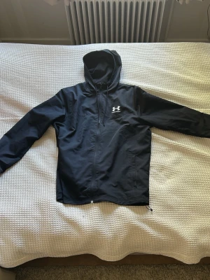 Svart vindjacka från Under Armour - Snygg svart vindjacka från Under Armour med dragkedja framtill, huva med dragsko och vit logga på bröstet. Jackan har långa ärmar och praktiska fickor. Perfekt för blåsiga dagar och sportiga outfits.
