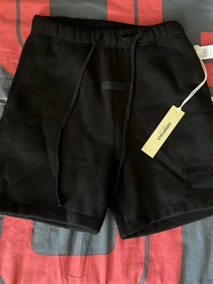 Svarta shorts från Essentials - Svarta shorts från Essentials med snörning i midjan och broderad logga på ena benet. De har en enkel och clean design, perfekt för en avslappnad stil. Tillverkade i mjukt material som känns skönt mot huden.