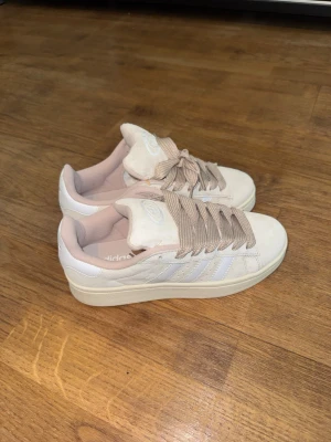 Vita Adidas sneakers med beige detaljer - Snygga vita Adidas sneakers med klassiska tre ränder på sidan och breda beigea skosnören. Skorna har en låg profil och diskret Adidas-logga på plösen. Insidan är ljusrosa och materialet är en mix av mocka och syntet. Perfekta för en clean och stilren look.