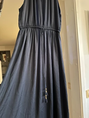 Svart ärmlös jumpsuit med snörning - Snygg svart ärmlös jumpsuit med markerad midja och snörningsdetalj med små guldfärgade pärlor. Jumpsuiten har vida ben och ett mjukt, följsamt tyg som ger en elegant look.