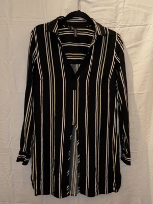 Svart-vit randig skjorta med knyt - Snygg långärmad skjorta med svart bas och vita vertikala ränder. Skjortan har klassisk krage, v-ringning och kan knytas framtill för en trendig look. Perfekt till jeans eller kjol för en cool stil.