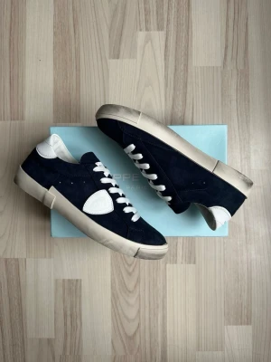 Mörkblå sneakers från Philippe Model - Snygga mörkblå sneakers från Philippe Model i mocka med vita detaljer och vit sula. Skorna har vita skosnören och en unik vit detalj på sidan samt vid hälen. Perfekta för dig som gillar stilrena och moderna sneakers med en twist.