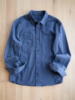 Navy overshirt i bomull - Säljer en stilren mörkblå overshirt i bomull med två stora bröstfickor och knappar framtill. Skjortan har klassisk krage och långa ärmar, perfekt att bära öppen eller stängd för en avslappnad look.