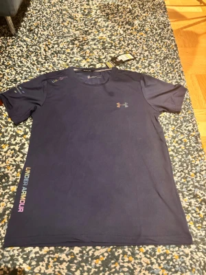 Blå tränings-tshirt från Under Armour - Snygg blå tränings-tshirt från Under Armour med rund hals och korta ärmar. Tillverkad i lätt och snabbtorkande HeatGear-material. Logga på bröstet och färgglad UNDER ARMOUR-text på sidan. Perfekt för löpning och gym.