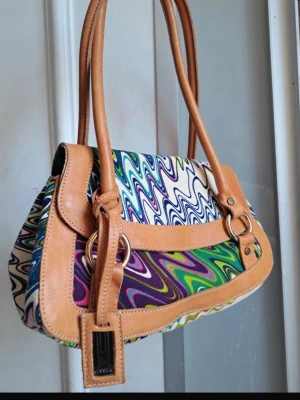 Mönstrad handväska från Missoni - Färgstark handväska från Missoni med vågigt mönster i lila, grönt, blått och vitt. Väskan har detaljer och handtag i ljusbrunt läder samt silverfärgade metalldelar. Insidan är ljus med dragkedjefack och Missoni-logga. Passar perfekt till ett par linnebyxor eller isabel marant skor. Perfekt för dig som gillar statement-accessoarer.                Bredd:25-30cm Höjd:15cm Djup:10cm