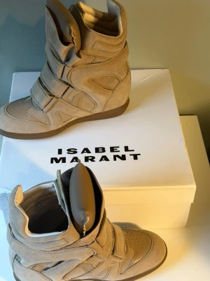 Isabel Marant Beige sneakers  - Snygga beige sneakers från Isabel Marant i mocka med högt skaft och tre kardborreband. Skorna har en chunky sula och cool design med paneldetaljer och vadderad plös. Perfekta för dig som vill ha en trendig och unik look.