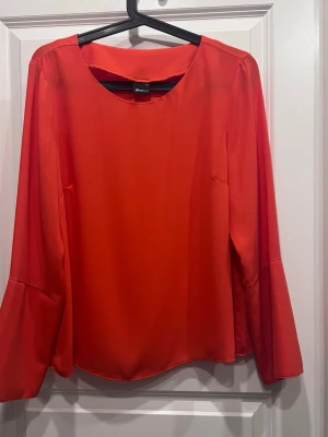 Röd/orange blus med vida ärmar. - Snygg röd/orange blus i ett glansigt, mjukt material med rund halsringning och långa, vida ärmar. Ärmarna har dekorativa knappar vid mudden och en liten slits. Perfekt för dig som vill sticka ut med färg och stil.