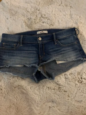 Blå jeansshorts från Hollister - Säljer ett par blå jeansshorts från Hollister med låg midja och fransiga kanter. Klassisk femficksmodell i mjuk denim. Perfekta till sommaren och ger en avslappnad vibe. 