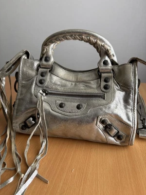 Silvrig handväska med nitar och fransar - Cool handväska i metallic-silver med svarta nitar och dragkedjor. Väskan har flätat handtag, dekorativa fransar och flera fack med dragkedja. Tillverkad i glansigt läderliknande material och har en unik, edgy design som sticker ut.