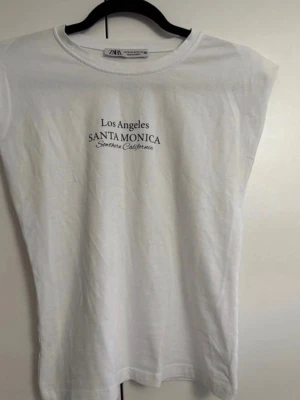 Vit ärmlös t-shirt från Zara - Snygg vit ärmlös t-shirt från Zara med tryck på bröstet där det står 'Los Angeles SANTA MONICA Southern California'. Perfekt till en avslappnad look och enkel att matcha med jeans eller shorts.