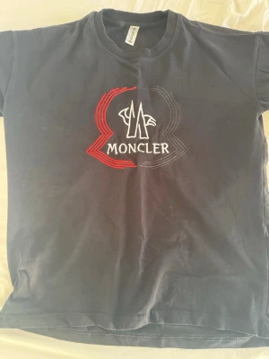 T shirt - Snygg svart t-shirt med stor broderad logga i vitt, rött och grått på bröstet. Klassisk rund hals och korta ärmar. Perfekt för dig som gillar streetwear och vill ha något stilrent men ändå med en twist. Storlek xl men passar som L