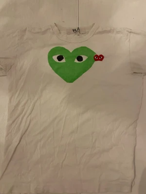 Vit Comme des Garçons Play t-shirt - Säljer en vit t-shirt från Comme des Garçons Play med det ikoniska gröna hjärtat med ögon och en liten röd detalj på bröstet. Klassisk passform och korta ärmar, perfekt till streetwear-looken.