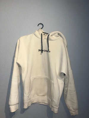 Vit hoodie från Palm Angels - Säljer en vit hoodie från Palm Angels med svart logga på bröstet och stor text över ryggen. Hoodien har huva med mörka snören och en stor magficka. Tillverkad i mjukt material som känns skönt mot huden. Perfekt för en avslappnad streetwear-look. Kom med bud!