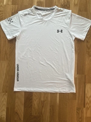Vit träningströja från Under Armour - Snygg vit t-shirt från Under Armour med logga på bröstet och tryck på ärmen samt längs sidan. Tillverkad i ett lätt och snabbtorkande material, perfekt för träning eller sport. Klassisk rund hals och korta ärmar för maximal rörelsefrihet.