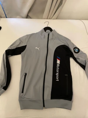 Puma BMW M Motorsport ziptröja grå - Snygg grå ziptröja från Puma med svart panel och BMW M Motorsport-logga på bröstet. Tröjan har hög krage, dragkedja framtill, två fickor med dragkedja och broderad Puma-logo. Perfekt för dig som gillar sportig stil och bilar.