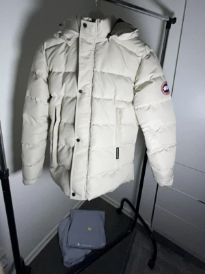 Vit dunjacka från Canada Goose - Snygg vit dunjacka från Canada Goose med klassisk patch på ärmen. Jackan har huva, stora fickor framtill och quiltad design som håller värmen. Perfekt för kalla vinterdagar och har en stilren look som passar till det mesta.