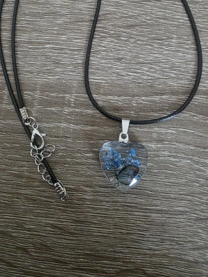 Halsband med hjärthänge och blå blommor - Unikt halsband med svart läderrem och ett hjärtformat hänge i transparent glas. Inuti hänget finns små blå blommor och en fjäril som ger en naturlig och konstnärlig känsla. Hänget har en metallögla och justerbart spänne i silverfärg.