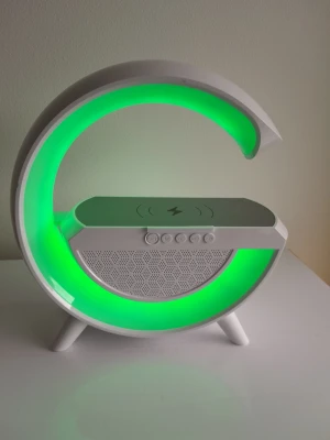 Ny BT 2301 G-formad LED trådlös laddare och Bluetooth-högtalare med FM-radio - Denna helt nya och multifunktionella BT 2301 LED-högtalare med trådlös laddning har en modern och stilren G-formad design som passar perfekt i sovrummet, på kontoret eller i arbetsrummet. Den är fullpackad med mångsidiga funktioner och fungerar som en allt-i-ett-lösning för stämningsbelysning, trådlös laddning och bärbart ljud. ​Trådlös laddningsplatta: Den platta ovansidan har en inbyggd trådlös laddningsplatta (markerad med en blixtsymbol) för att snabbt och smidigt ladda din smartphone. ​Dyna