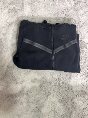 Svart Nike hoodie med dragkedja (147-158cm) - Säljer en svart hoodie från Nike med dragkedja framtill och en snygg grå detalj över bröstet. Hoodien har huva och Nike-logga på vänster sida. Perfekt för en sportig och avslappnad stil.