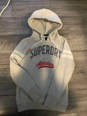 Ljusgrå hoodie från Superdry XS - Ljusgrå hoodie från Superdry med stor huva och trycket 'Superdry Athletics' i mörkblått och rött på bröstet. Två vita ränder på ärmarna ger en sportig vibe. Mjuk insida och breda muddar vid ärmslut och nederkant. Perfekt för chill dagar.