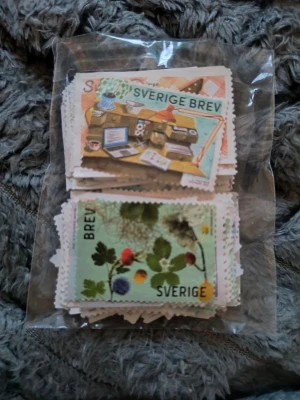 Svenska frimärken i plastpåse - 100st ostämplade Sverige brev utan lim. Alla är fina. Ett gäng svenska frimärken i olika motiv, bland annat med natur och skrivbordstema. Frimärkena är rektangulära med tandade kanter och färgglada illustrationer. De ligger samlade i en genomskinlig plastpåse. Perfekt för samlare eller pyssel.