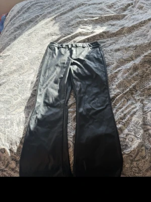 Svarta utsvängda byxor i skinnimitation - Säljer ett par svarta byxor i skinnimitation med utsvängda ben och hög midja. Snyggt glansigt material som ger en cool look. Perfekta för dig som vill sticka ut lite extra. Passar till många olika stilar!