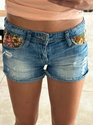 Lågmidjade blå jeansshorts med blommiga detaljer - Säljer ett par lågmidjade blå jeansshorts med slitna detaljer och blommigt tyg på fickorna fram och bak. Shortsen har upprullade benslut och klassisk femficksdesign. Perfekta för sommaren och ger en cool vintagekänsla. Nyspriset är cirka 350 och säljer för 250!!💕 